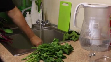 Bir adam, mutfaktaki bir su deresinin altında güçlü bir şekilde bir miktar kereviz yıkıyor. Sonra suyu silkeliyor ve kerevizi lavabonun sağına koyuyor..