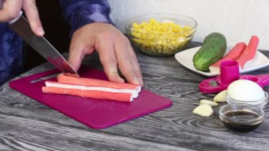 Bir adam kesme tahtasındaki yengeç çubuklarını kesiyor. Salata hazırlamak için yengeç çubukları, tavuk yumurtası ve sebze. Boyalı çam ağaçlarında yatıyorlar..