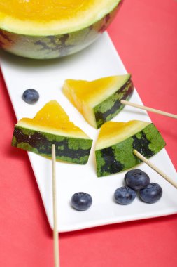 Çubuklardaki sarı karpuz parçaları. Tabağa uzan. Yakınlarda yabanmersini var. Mercan renkli bir yüzeyde bir tabak..