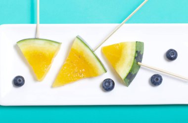 Çubuklardaki sarı karpuz parçaları. Tabağa uzan. Yakınlarda yabanmersini var. Nane renginde bir yüzey üzerindeki tabaka.