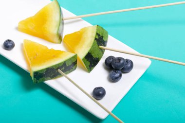 Çubuklardaki sarı karpuz parçaları. Tabağa uzan. Yakınlarda yabanmersini var. Nane renginde bir yüzey üzerindeki tabaka.