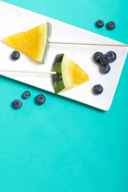 Çubuklardaki sarı karpuz parçaları. Tabağa uzan. Yakınlarda yabanmersini var. Nane renginde bir yüzey üzerindeki tabaka.