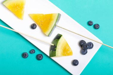 Çubuklardaki sarı karpuz parçaları. Tabağa uzan. Yakınlarda yabanmersini var. Nane renginde bir yüzey üzerindeki tabaka.