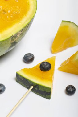 Çubuklardaki sarı karpuz parçaları. Yakınlarda yabanmersini var. Beyaz bir yüzeyde plaka.