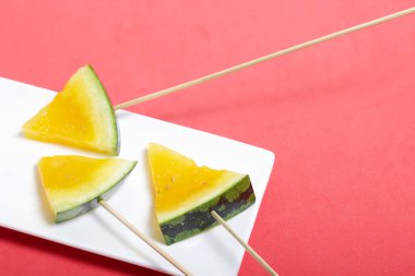 Çubuklardaki sarı karpuz parçaları. Tabağa uzan. Mercan renkli bir yüzeyde bir tabak..