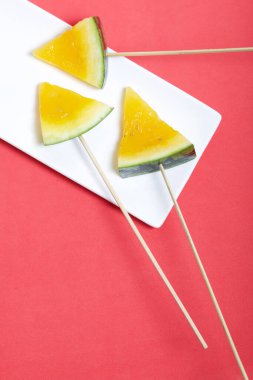 Çubuklardaki sarı karpuz parçaları. Tabağa uzan. Mercan renkli bir yüzeyde bir tabak..