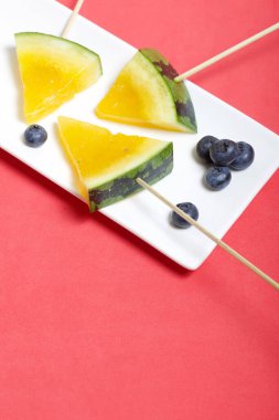 Çubuklardaki sarı karpuz parçaları. Tabağa uzan. Yakınlarda yabanmersini var. Mercan renkli bir yüzeyde bir tabak..