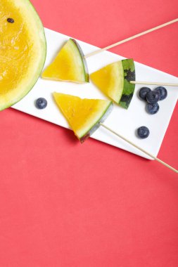 Çubuklardaki sarı karpuz parçaları. Tabağa uzan. Yakınlarda yabanmersini var. Mercan renkli bir yüzeyde bir tabak..