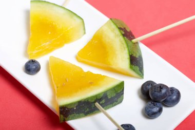 Çubuklardaki sarı karpuz parçaları. Tabağa uzan. Yakınlarda yabanmersini var. Mercan renkli bir yüzeyde bir tabak..