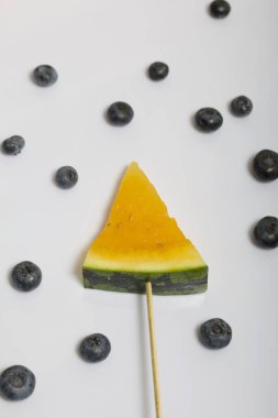 Çubuklardaki sarı karpuz parçaları. Yakınlarda yabanmersini var. Beyaz bir yüzeyde plaka.