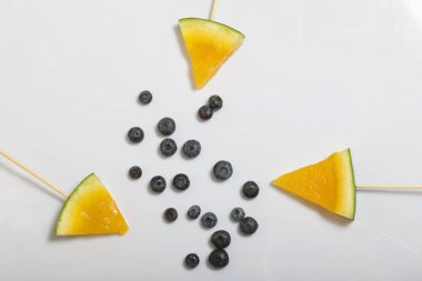 Çubuklardaki sarı karpuz parçaları. Yakınlarda yabanmersini var. Beyaz bir yüzeyde plaka.