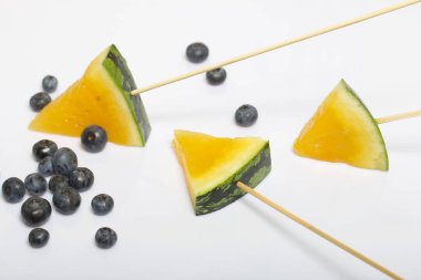 Çubuklardaki sarı karpuz parçaları. Yakınlarda yabanmersini var. Beyaz bir yüzeyde plaka.