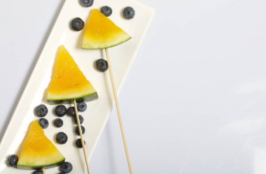 Çubuklardaki sarı karpuz parçaları. Tabağa uzan. Yakınlarda yabanmersini var. Beyaz bir yüzeyde plaka.