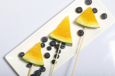 Çubuklardaki sarı karpuz parçaları. Tabağa uzan. Yakınlarda yabanmersini var. Beyaz bir yüzeyde plaka.