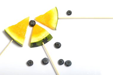 Çubuklardaki sarı karpuz parçaları. Yakınlarda yabanmersini var. Beyaz bir yüzeyde plaka.