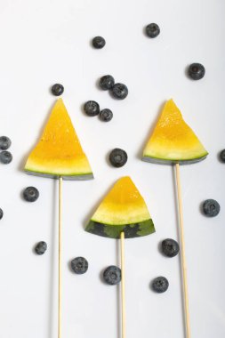 Çubuklardaki sarı karpuz parçaları. Yakınlarda yabanmersini var. Beyaz bir yüzeyde plaka.