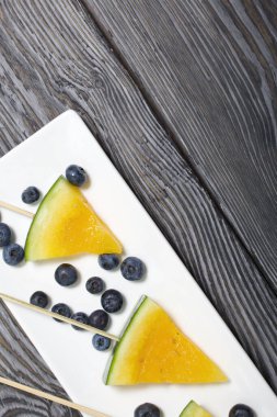 Çubuklardaki sarı karpuz parçaları. Tabağa uzan. Yakınlarda yabanmersini var. Tabak boyalı çam ağaçlarında.