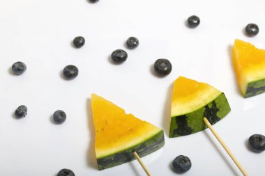 Çubuklardaki sarı karpuz parçaları. Yakınlarda yabanmersini var. Beyaz bir yüzeyde plaka.