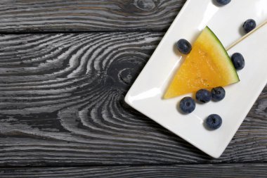 Çubuklardaki sarı karpuz parçaları. Tabağa uzan. Yakınlarda yabanmersini var. Tabak boyalı çam ağaçlarında.