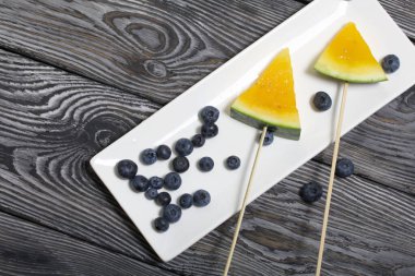 Çubuklardaki sarı karpuz parçaları. Tabağa uzan. Yakınlarda yabanmersini var. Tabak boyalı çam ağaçlarında.