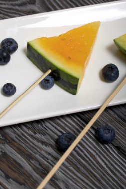 Çubuklardaki sarı karpuz parçaları. Tabağa uzan. Yakınlarda yabanmersini var. Tabak boyalı çam ağaçlarında.