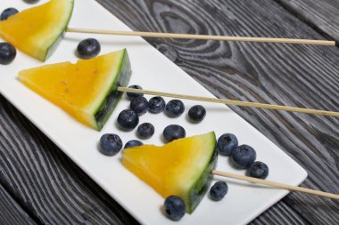 Çubuklardaki sarı karpuz parçaları. Tabağa uzan. Yakınlarda yabanmersini var. Tabak boyalı çam ağaçlarında.