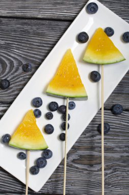 Çubuklardaki sarı karpuz parçaları. Tabağa uzan. Yakınlarda yabanmersini var. Tabak boyalı çam ağaçlarında.