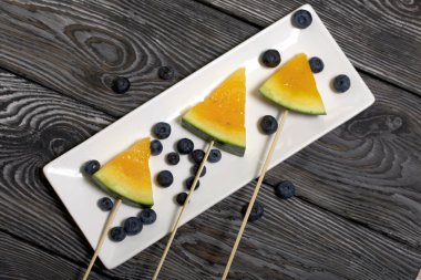 Çubuklardaki sarı karpuz parçaları. Tabağa uzan. Yakınlarda yabanmersini var. Tabak boyalı çam ağaçlarında.