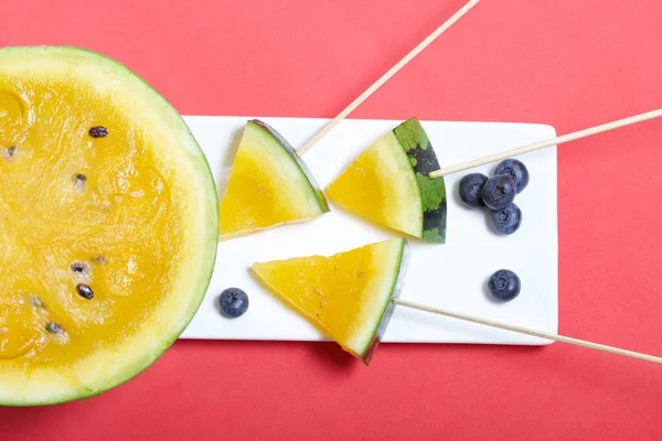 Çubuklardaki sarı karpuz parçaları. Tabağa uzan. Yakınlarda yabanmersini var. Mercan renkli bir yüzeyde bir tabak..