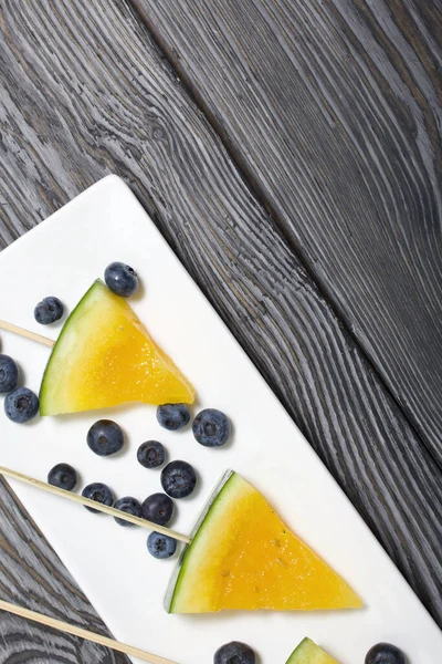 Çubuklardaki sarı karpuz parçaları. Tabağa uzan. Yakınlarda yabanmersini var. Tabak boyalı çam ağaçlarında.