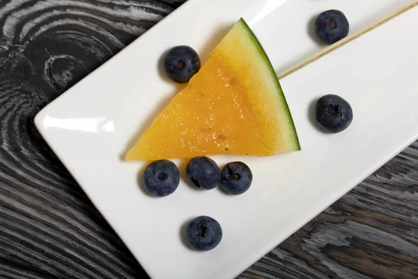 Çubuklardaki sarı karpuz parçaları. Tabağa uzan. Yakınlarda yabanmersini var. Tabak boyalı çam ağaçlarında.