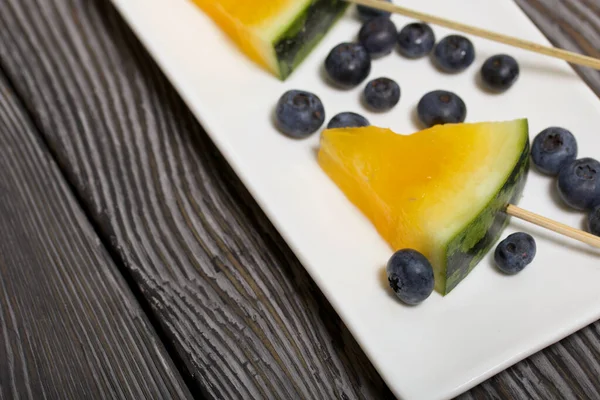 Çubuklardaki sarı karpuz parçaları. Tabağa uzan. Yakınlarda yabanmersini var. Tabak boyalı çam ağaçlarında.