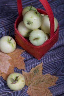 Kırmızı hasır sepetteki elmalar. Yakınlarda elmalar ve kurumuş akçaağaç yaprakları var. Meyve hasadı.