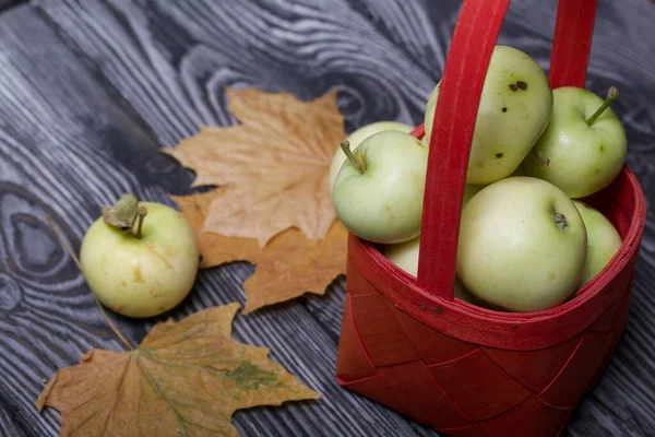 Kırmızı hasır sepetteki elmalar. Yakınlarda elmalar ve kurumuş akçaağaç yaprakları var. Meyve hasadı.