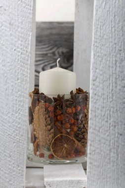 Cam şamdanda bir mum. Şamdan kurutulmuş koniler, baharatlar ve kurutulmuş meyvelerle dolu. Tahta bir kutunun içinde duruyor..