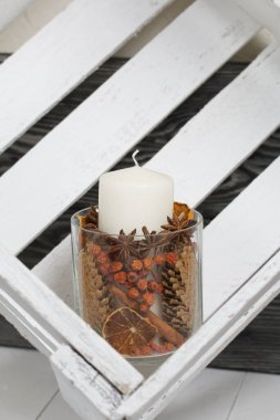 Cam şamdanda bir mum. Şamdan kurutulmuş koniler, baharatlar ve kurutulmuş meyvelerle dolu. Tahta bir kutunun içinde duruyor..