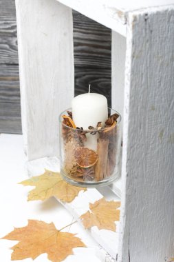 Cam şamdanda bir mum. Şamdan kurutulmuş koniler, baharatlar ve kurutulmuş meyvelerle dolu. Tahta bir kutunun içinde duruyor. Yakınlarda kurumuş akçaağaç yaprakları var..