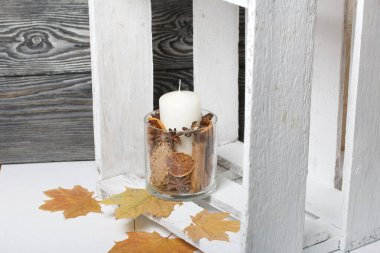 Cam şamdanda bir mum. Şamdan kurutulmuş koniler, baharatlar ve kurutulmuş meyvelerle dolu. Tahta bir kutunun içinde duruyor. Yakınlarda kurumuş akçaağaç yaprakları var..