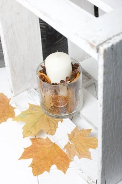 Cam şamdanda bir mum. Şamdan kurutulmuş koniler, baharatlar ve kurutulmuş meyvelerle dolu. Tahta bir kutunun içinde duruyor. Yakınlarda kurumuş akçaağaç yaprakları var..