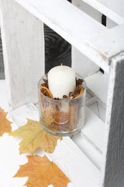 Cam şamdanda bir mum. Şamdan kurutulmuş koniler, baharatlar ve kurutulmuş meyvelerle dolu. Tahta bir kutunun içinde duruyor. Yakınlarda kurumuş akçaağaç yaprakları var..