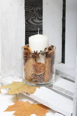 Cam şamdanda bir mum. Şamdan kurutulmuş koniler, baharatlar ve kurutulmuş meyvelerle dolu. Tahta bir kutunun içinde duruyor. Yakınlarda kurumuş akçaağaç yaprakları var..