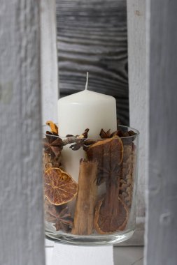Cam şamdanda bir mum. Şamdan kurutulmuş koniler, baharatlar ve kurutulmuş meyvelerle dolu. Tahta bir kutunun içinde duruyor..