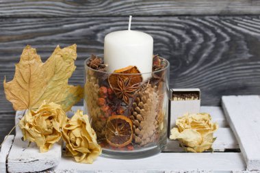 Cam şamdanda bir mum. Şamdan kurutulmuş koniler, baharatlar ve kurutulmuş meyvelerle dolu. Tahta bir kutunun üzerinde duruyor. Yakınlarda kurumuş güller ve akçaağaç yaprakları var..