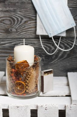 Cam şamdanda bir mum. Şamdan kurutulmuş koniler, baharatlar ve kurutulmuş meyvelerle dolu. Tahta bir kutunun üzerinde duruyor. Tıbbi maskeler yakınlarda asılı..