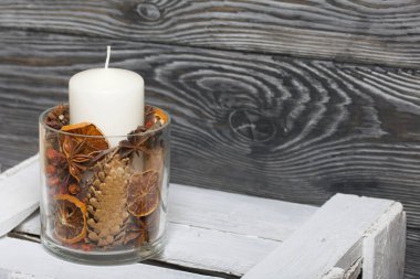 Cam şamdanda bir mum. Şamdan kurutulmuş koniler, baharatlar ve kurutulmuş meyvelerle dolu. Tahta bir kutunun üzerinde duruyor..