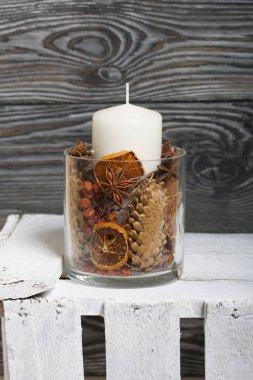 Cam şamdanda bir mum. Şamdan kurutulmuş koniler, baharatlar ve kurutulmuş meyvelerle dolu. Tahta bir kutunun üzerinde duruyor..