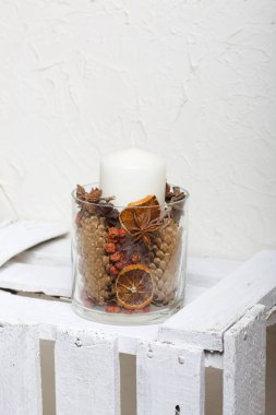 Cam şamdanda bir mum. Şamdan kurutulmuş koniler, baharatlar ve kurutulmuş meyvelerle dolu. Tahta bir kutunun üzerinde duruyor..
