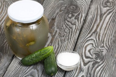 Bir kavanoz salatalık turşusu. Yakınlarda taze salatalıklar ve bir kap tuz var. Boyalı bir tahta yüzeyde.