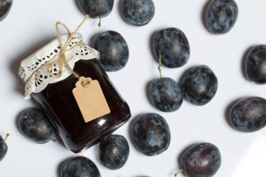 Bir kavanoz erik reçeli. Üzerinde bir yazıt etiketi var. Erikler yakınlarda dağılmış. Beyaz bir arkaplanda.