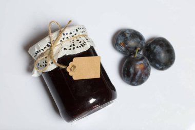 Bir kavanoz erik reçeli. Üzerinde bir yazıt etiketi var. Erikler yakınlarda dağılmış. Beyaz bir arkaplanda.